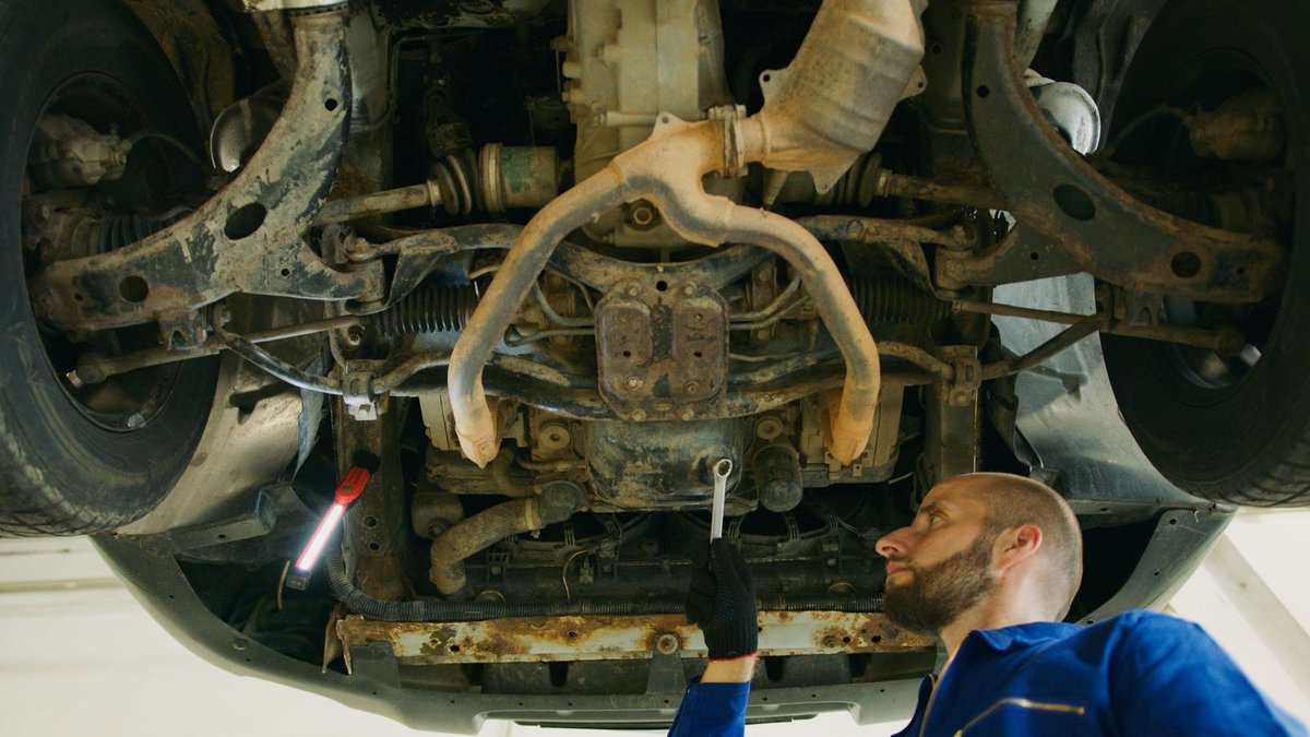 Mechanic with a wrench inspecting the underside of a lifted car, with the CV axles, control arms, and exhaust visible overhead