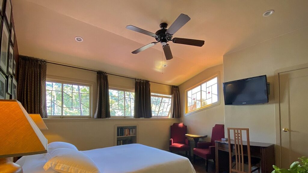 A dark five-blade ceiling fan on the ceiling of a bright, sunlit bedroom with a white bed and warm lamp light