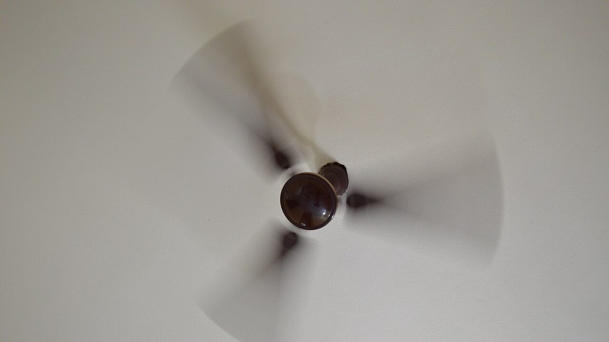 A brown ceiling fan photographed from directly below while spinning, its blades blurred with motion against a white ceiling
