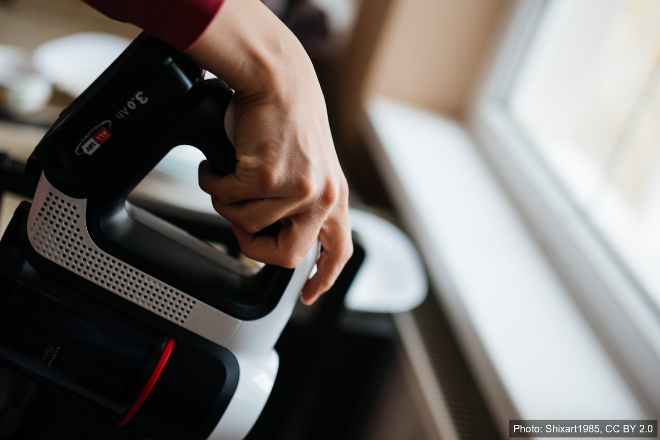 A hand gripping a real cordless stick vacuum by a window, deep-cleaning by hand where a robot vacuum cannot reach