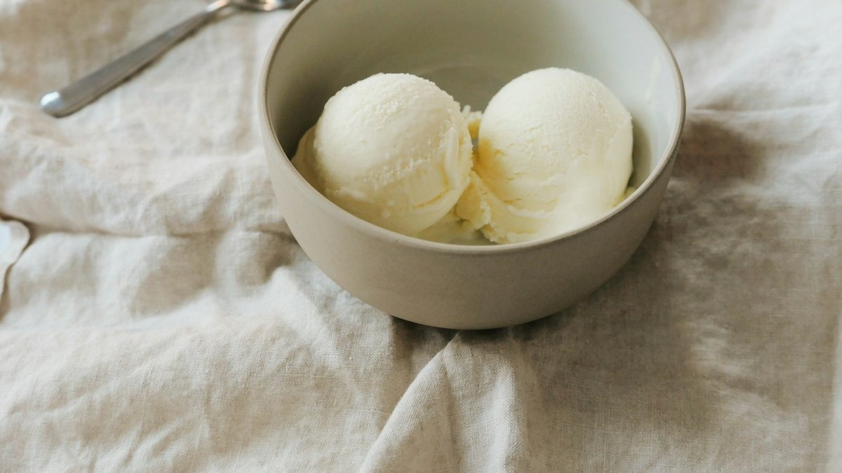 Two scoops of pale, creamy homemade cottage cheese ice cream in a ceramic bowl with a spoon resting beside it