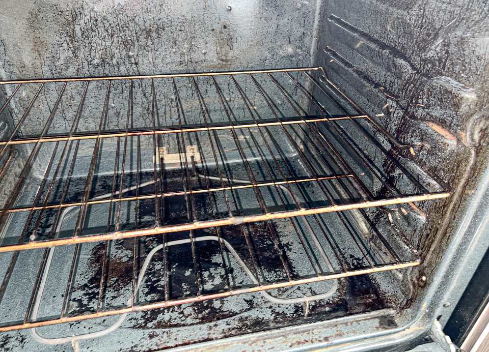 The filthy interior of a neglected oven, its floor and walls coated in thick black baked-on grease, with two greasy wire racks still inside