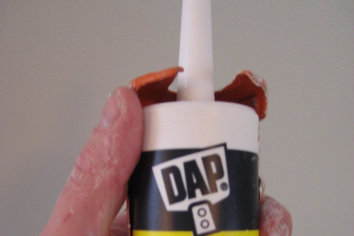 A hand holding a tube of DAP acrylic latex caulk with the nozzle ready to seal a gap