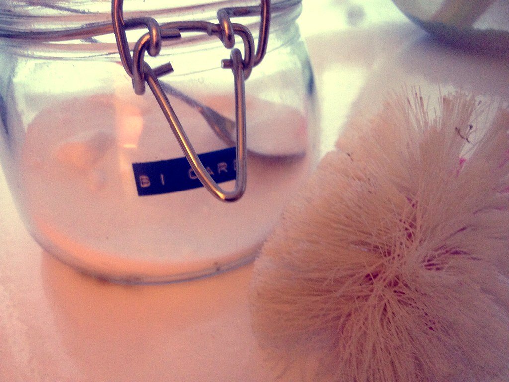 A glass jar of bicarbonate of soda (baking soda) next to a natural-bristle scrub brush on a countertop