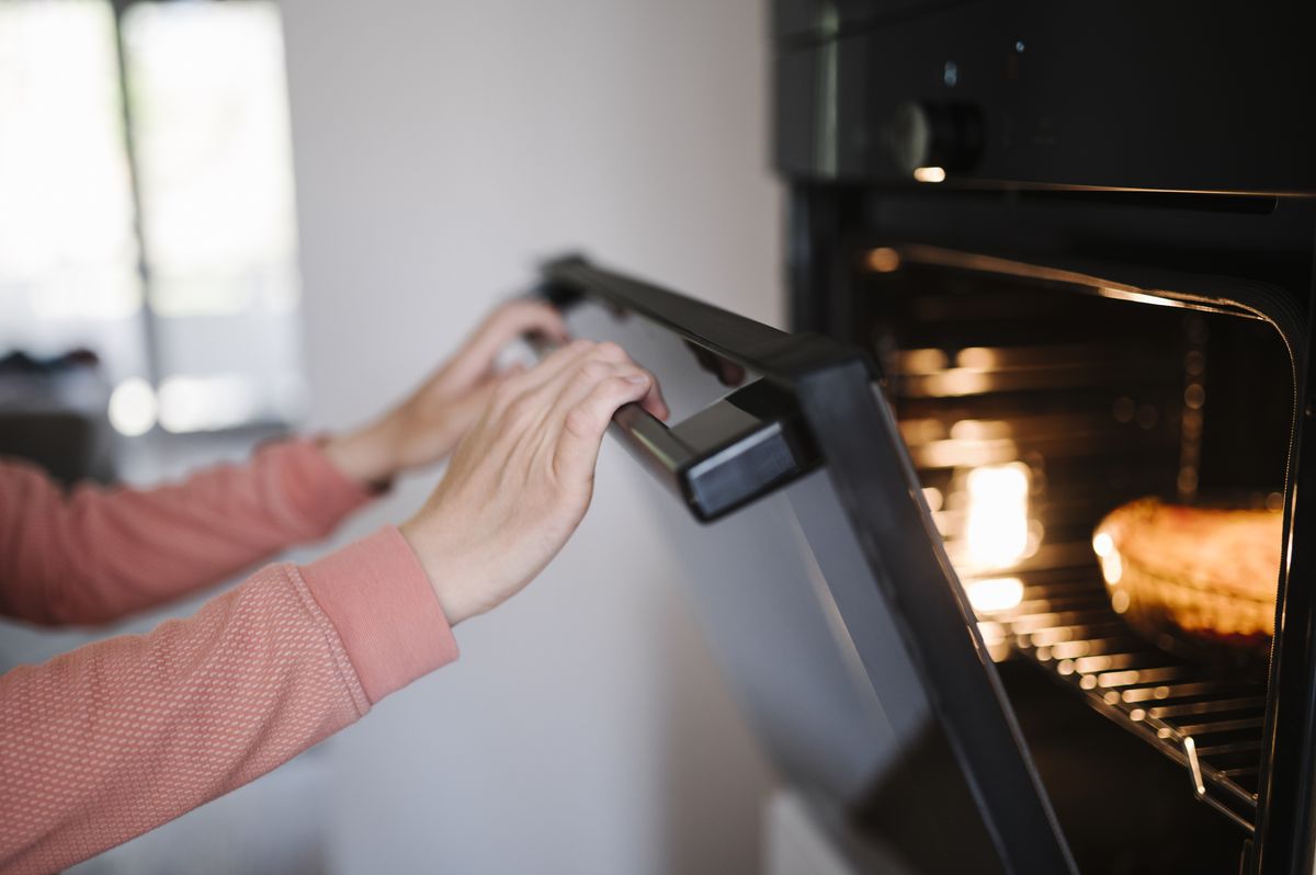 Hands opening the door of a modern black wall oven with a control knob at the top and food glowing on the rack inside
