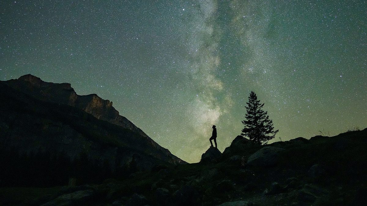 A skywatcher standing on a rock under the Milky Way while watching the Perseid meteor shower from a dark mountain site