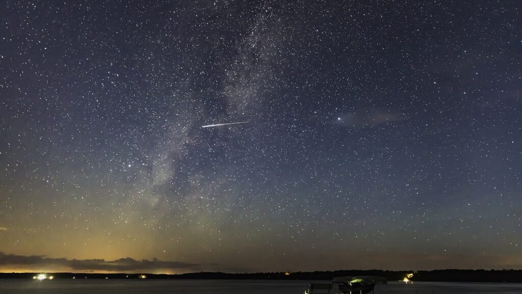 A Perseid meteor streaking across a dark star-filled sky and the Milky Way above a quiet lake on a moonless night