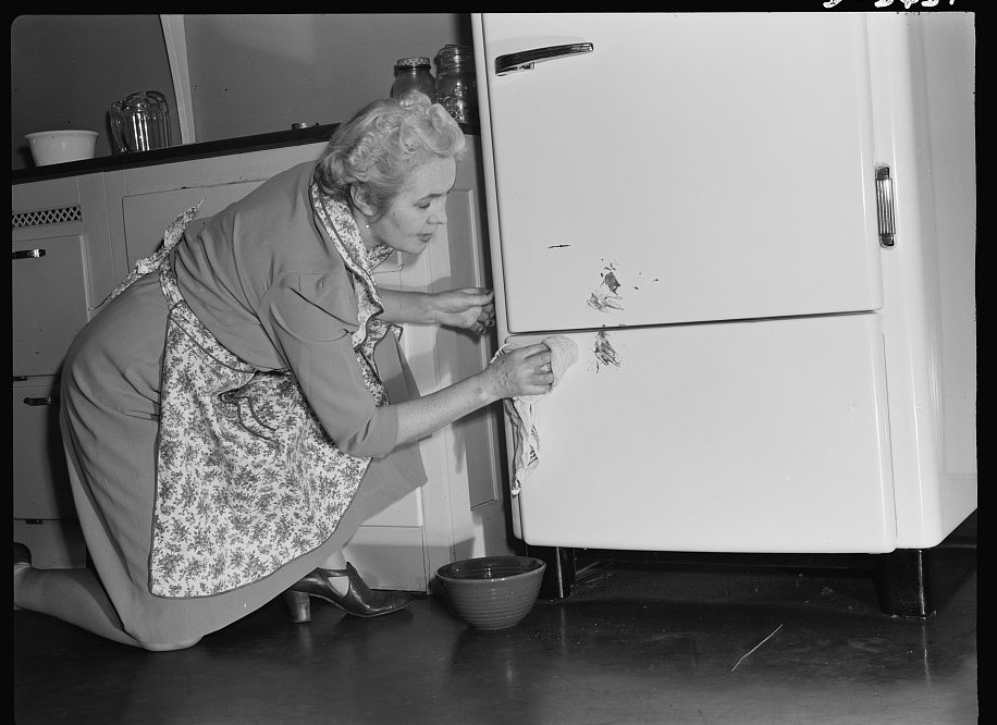 A person kneeling and wiping a spilled food stain off the front of a refrigerator with a damp cloth and a bowl of water