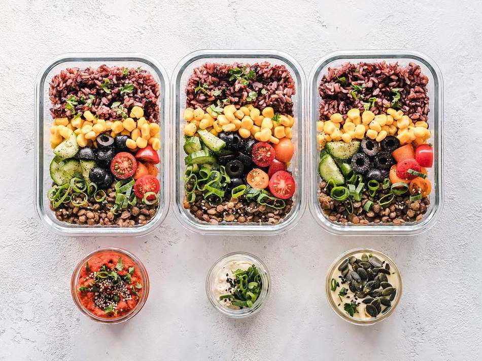 Three glass meal-prep containers with airtight lids full of rice, corn, vegetables and lentils, plus small sealed dip jars