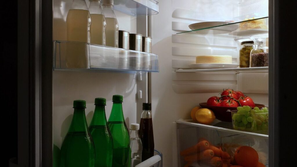 An open home refrigerator glowing at night, packed with bottles, cans, cheese, tomatoes, grapes, carrots and oranges on every shelf