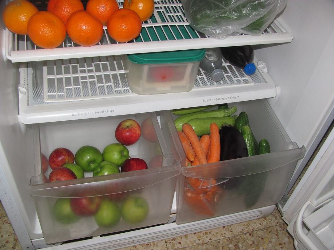 Open refrigerator crisper drawers pulled out, showing apples, carrots, zucchini and cucumbers where spoiled produce and old spills hide