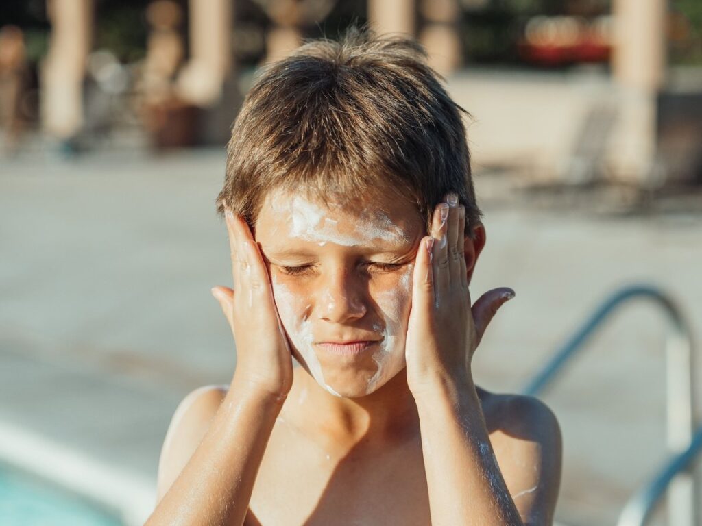 Close-up of a child pressing visible white sunscreen into his cheeks at poolside before it fully blends in