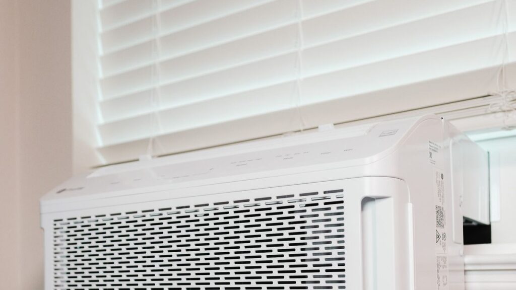 A white window-mounted air conditioner unit installed in a residential window with white blinds, seen from inside a room